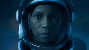 Kosmonauts baltā aizsargķiverē ar ļoti nopietnu sejas izteiksmi skatās tieši kameras objektīvā zilganā un dūmakainā apgaismojumā.