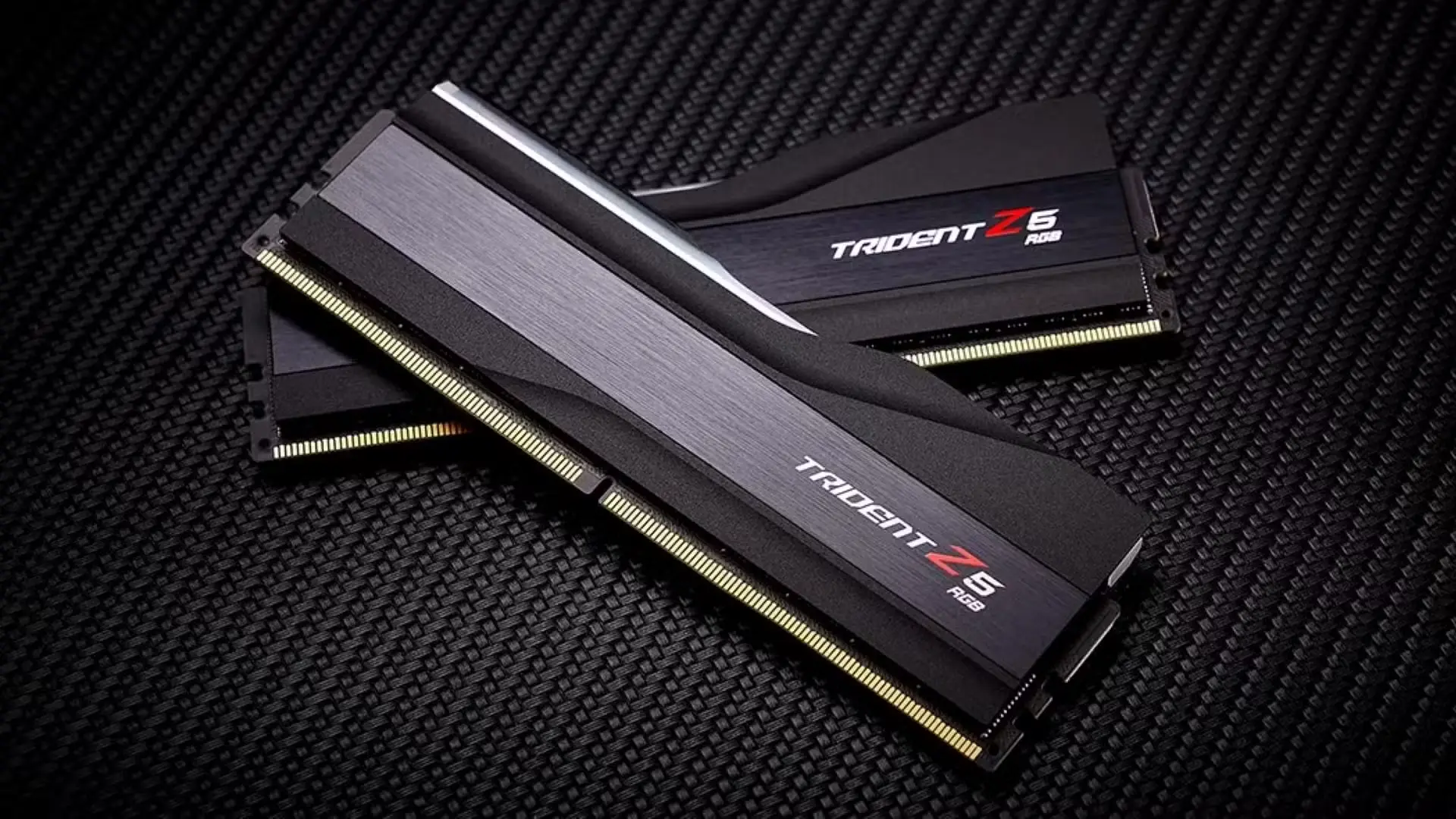 Divi melni G.Skill Trident Z5 DDR5 operatīvās atmiņas moduļi ar sudraba elementiem guļ uz oglekļa šķiedras virsmas.