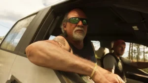 Vecāks vīrietis ar saulesbrillēm un bārdu skatās pa automašīnas logu, kamēr otrs vīrietis ar cepuri vada auto spēles GTA 6 reklāmas attēlā
