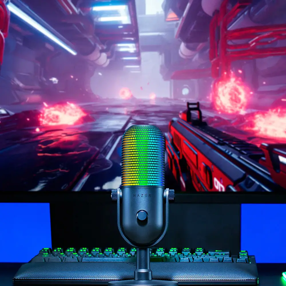 Razer Seiren V3 Chroma mikrofons uz spēlētāja galda pie monitora spraigas datorspēles laikā.