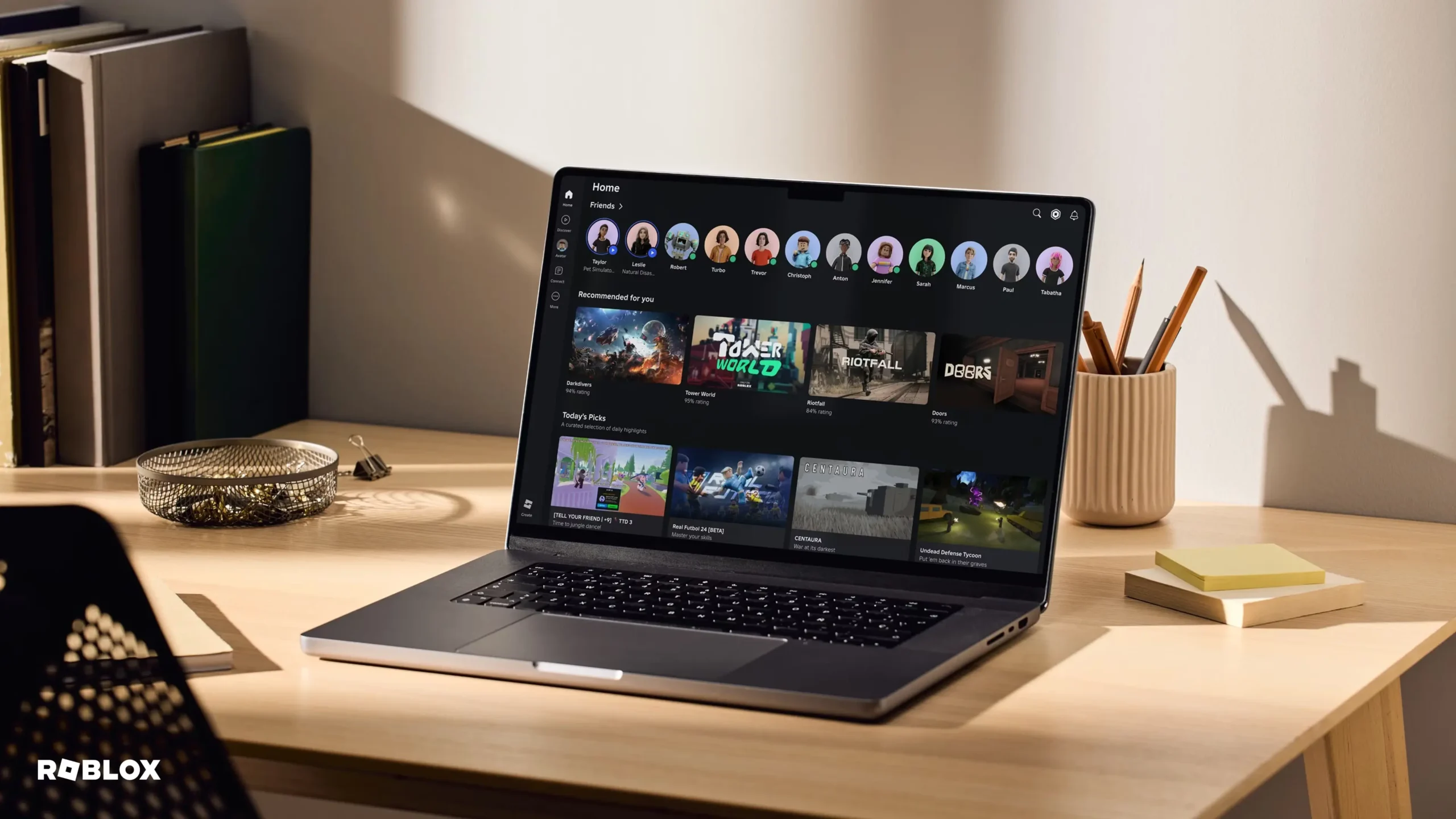 Klēpjdators atrodas uz koka galda gaišā istabā, un tā ekrānā ir redzama Roblox mājaslapa ar spēļu ieteikumiem un draugu sarakstu.