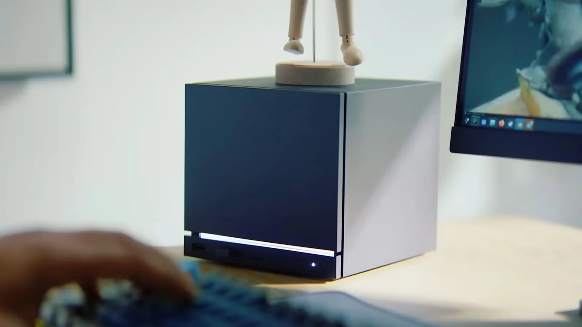 Pelēka un kubveida Valve Steam Machine mājas konsole atrodas uz gaiša koka galda blakus datora monitoram un klaviatūrai, kuras priekšplānā redzama roka.