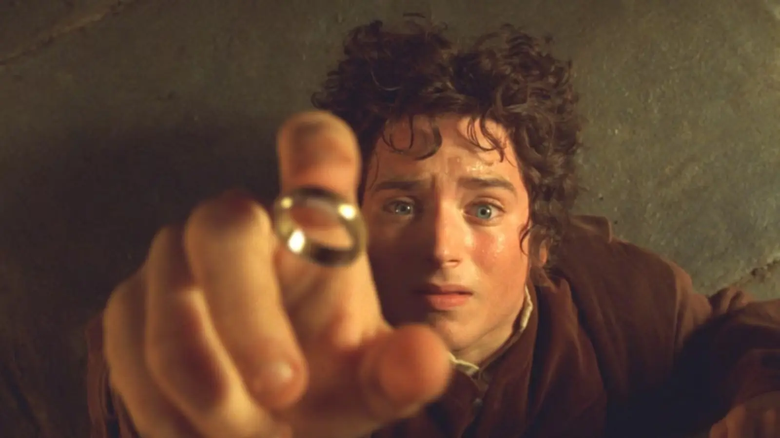 Frodo no apakšas skata mērķtiecīgi sniedzas ar roku uz priekšu, turot starp pirkstiem mirdzošu Viena Gredzena zelta gredzenu.