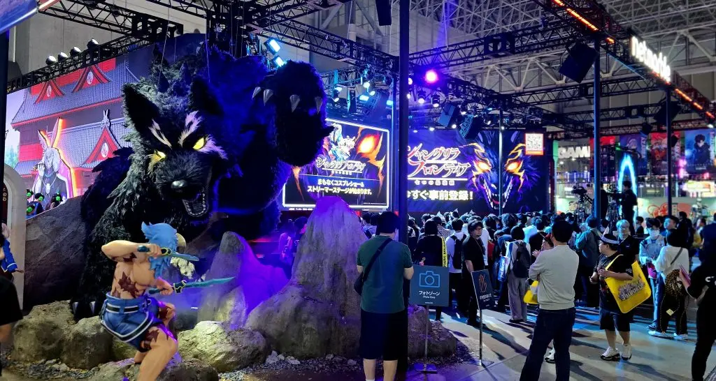 Milzīgs tumšs vilkveidīgs briesmonis un karavīra statuja Netmarble spēļu stendā Tokyo Game Show izstādē ar blīvu apmeklētāju pūli fonā