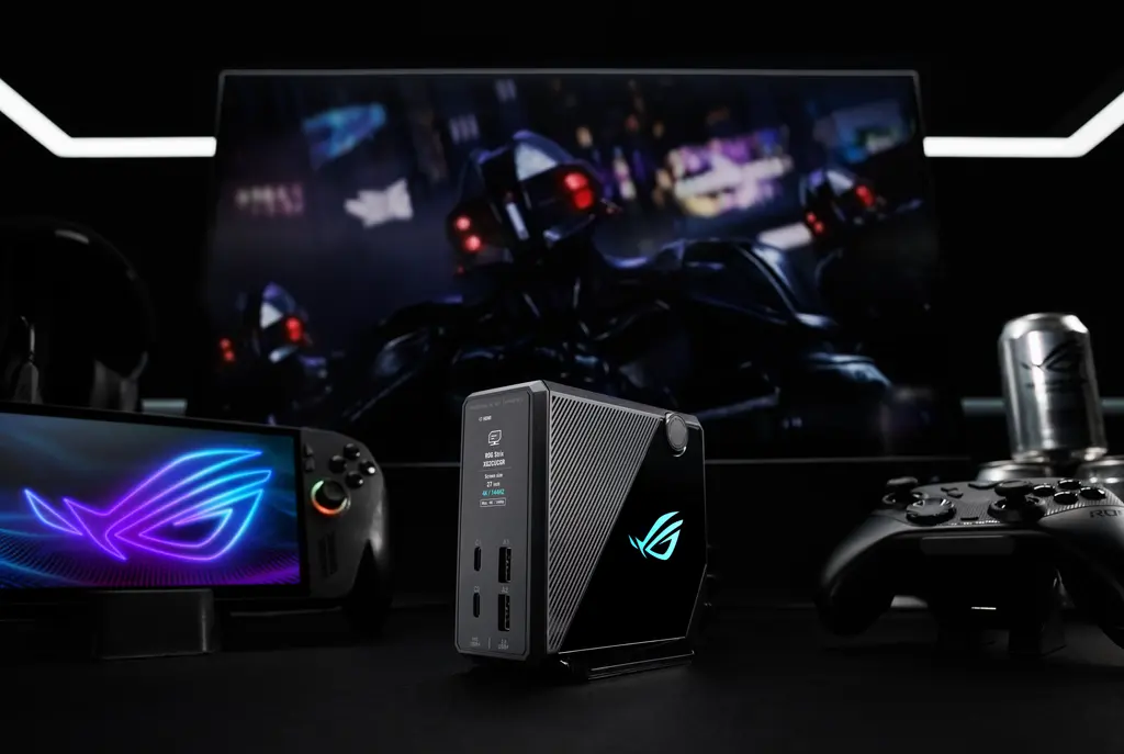 Melna ASUS ROG Flux Charger Dock uzlādes stacija ar zili izgaismotu logotipu un informācijas ekrānu, kas novietota uz galda blakus monitoram, spēļu pultij un pārnēsājamai konsolei