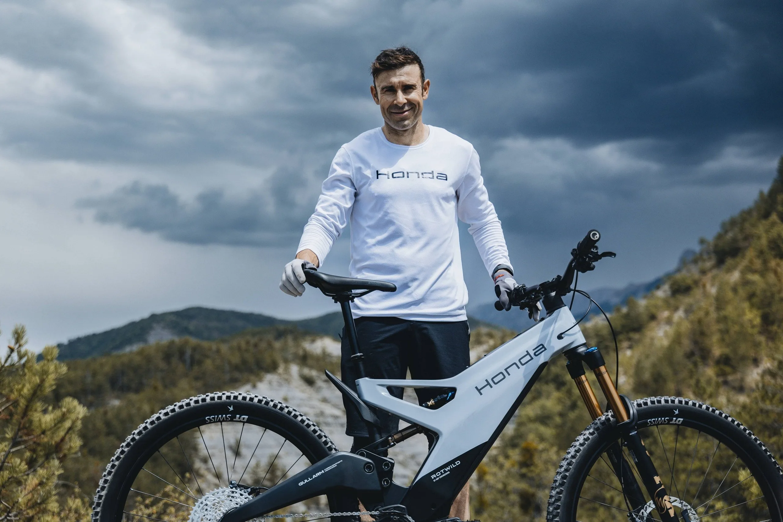 Tonijs Bou baltā Honda kreklā stāv blakus jaunam pelēkbaltam Honda elektriskajam kalnu velosipēdam kalnu un mākoņainu debesu fonā