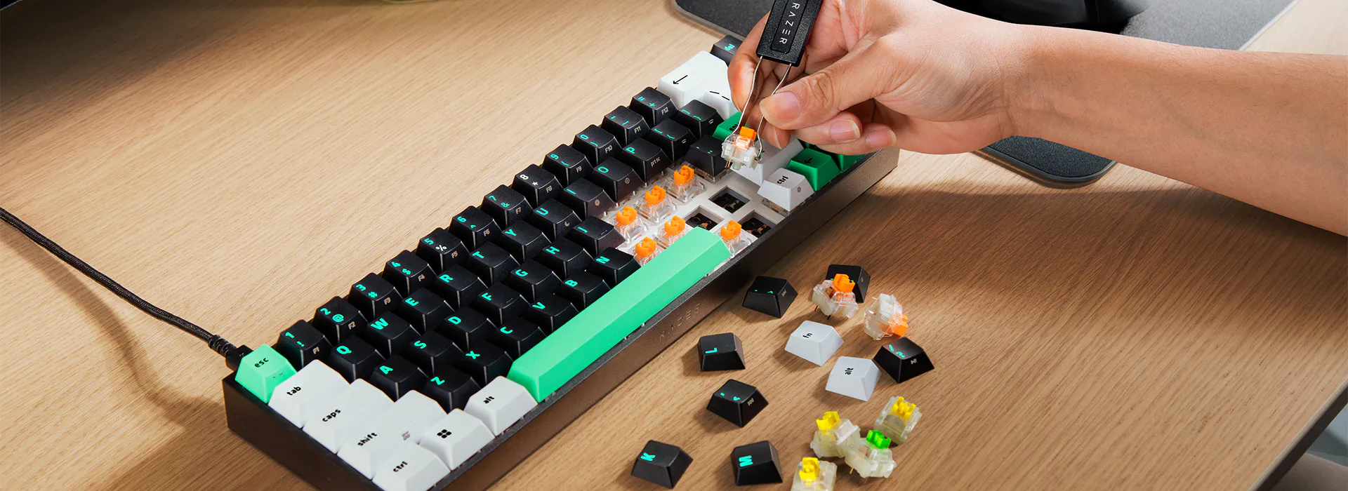 Lietotāja roka ar speciālu rīku izņem oranžu mehānisko slēdzi no Razer Reclusa X Mini 65% klaviatūras, kurai ir melni, balti un zaļi taustiņi.